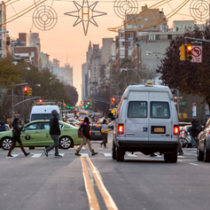 A white van is stopped at a red light on a busy intersection. Pedestrians cross on the crosswalk in front of the van.”