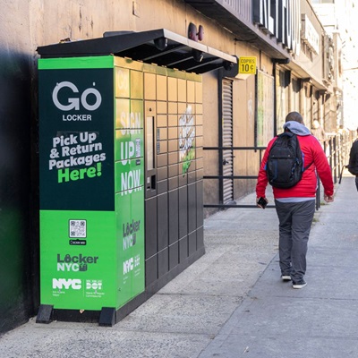 A person walks by a stack of green and black lockers with Go Locker Pick Up and Return Packages text on them, are positioned along a supermarket’s wall on a Brooklyn sidewalk.