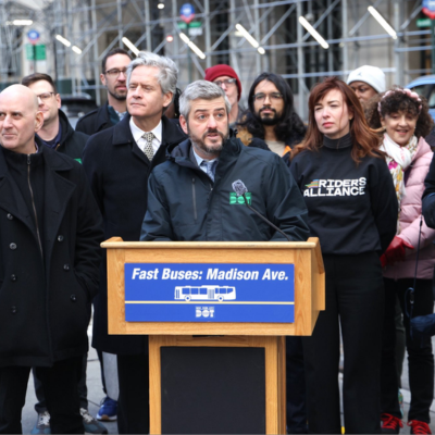 Department of Transportation Commissioner Mike Flynn stands behind a podium with multiple people standing around him. Podium signs reads 