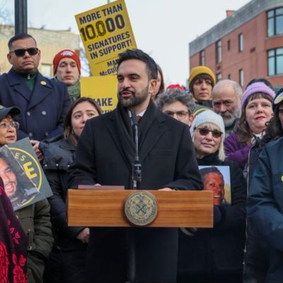 Mayor Mamdani stands behind a podium with multiple people standing around him.