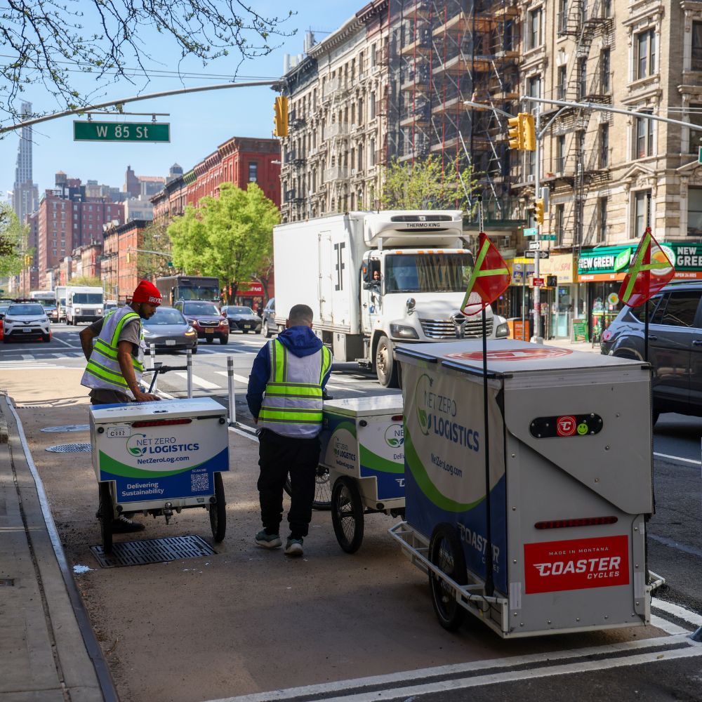Two delivery workers place packages in cargo bikes parked in a curb extension along a busy roadway.