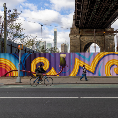 People walk and bike in front of a fence painted with a mural of a dandelion flower and other brightly colored shapes.