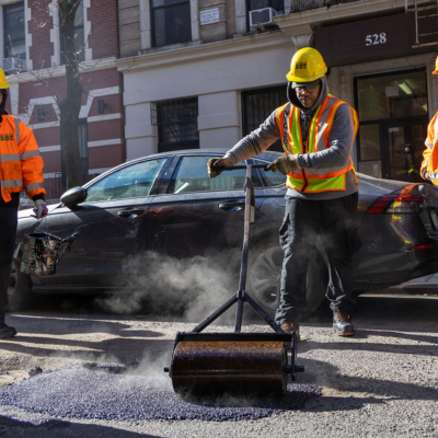 A D O T worker in high visibility gear rolls and compacts hot asphalt over a pothole on a city street.