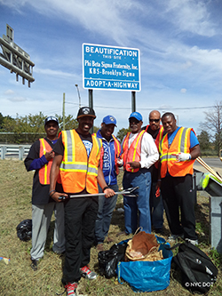 A group of men wearing orange vests stand in front of their Adopt a Highway sign. The sign reads Phi Beta Sigma Fraternity, Inc. K B S – Brooklyn Sigma 