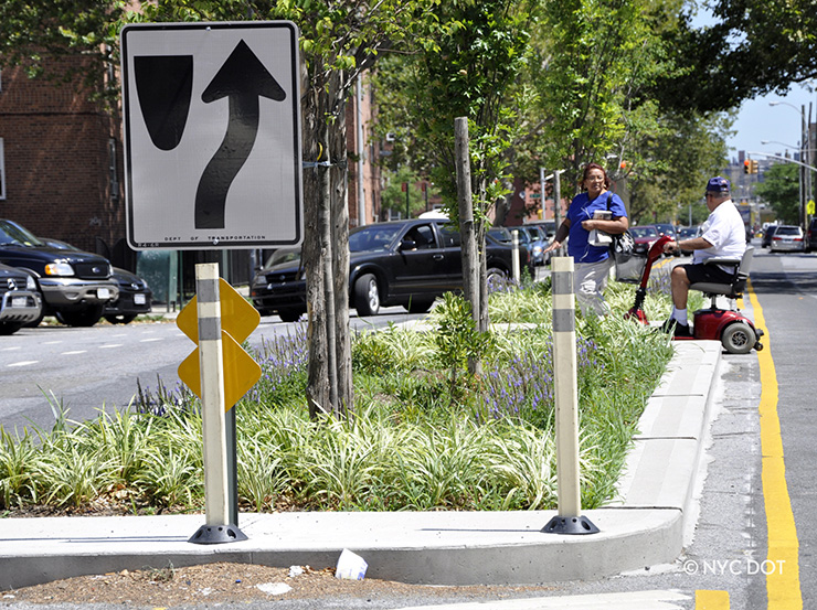 A pedestrian refuge island planted with trees in Red Hook, Brooklyn. Two pedestrians, on in a mobility scooter, are waiting to cross the street.