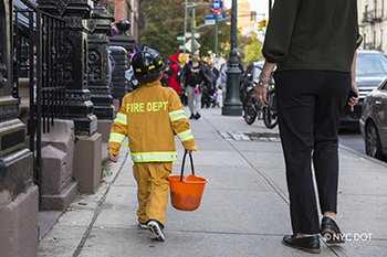 During a Halloween celebration in NYC, a young person dressed as a fire fighter walks on a wide sidewalk in front of townhouses. 
