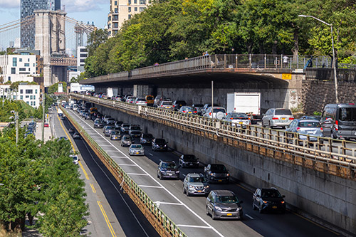 Dense car and truck traffic travels along the Brooklyn Queens Expressway’s three tiered roadway. Stacked above the roadway is a pedestrian promenade.