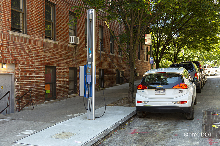 A white electric vehicle is parked along a curb in the Bronx in a special space reserved for charging electric vehicles. Installed on the sidewalk next to the parked car is a tall pole with “Flo” and “PlugNYC” branding. The pole has two long cables with plugs to charge electric vehicles parked in the curbside spaces.