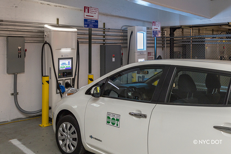 A white Department of Transportation vehicle is parked in a municipal garage and plugged into a DC fast charging station.