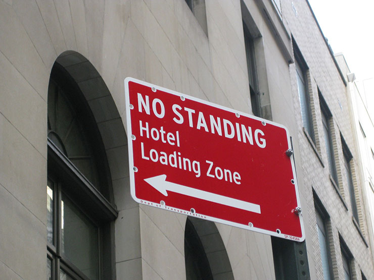 Red rectangular street sign with white text saying "No Standing Hotel Loading Zone" 