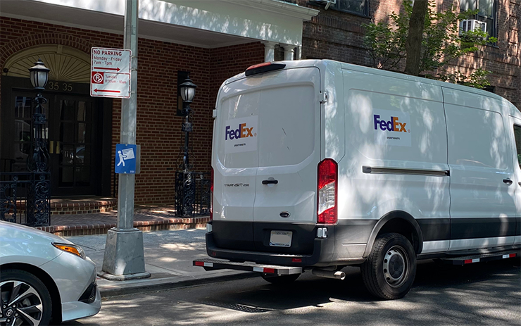 A white FedEx van parks along the curb in front of a residential building besides a No Parking Monday to Friday sign. Lower on the post is a blue Neighborhood Loading Zone sign.