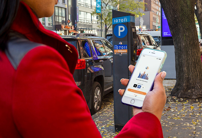 A woman stands on the sidewalk next to a parked car and a parking meter. She holds her phone and opens the Park N Y C app to pay for her parking.