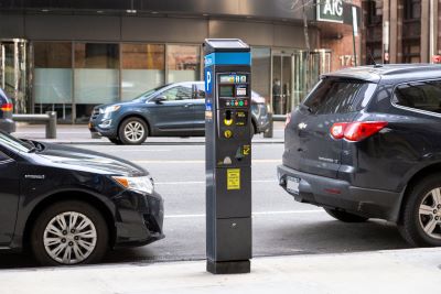 A parking meter is positioned on a sidewalk near a street with cars parked along the curb