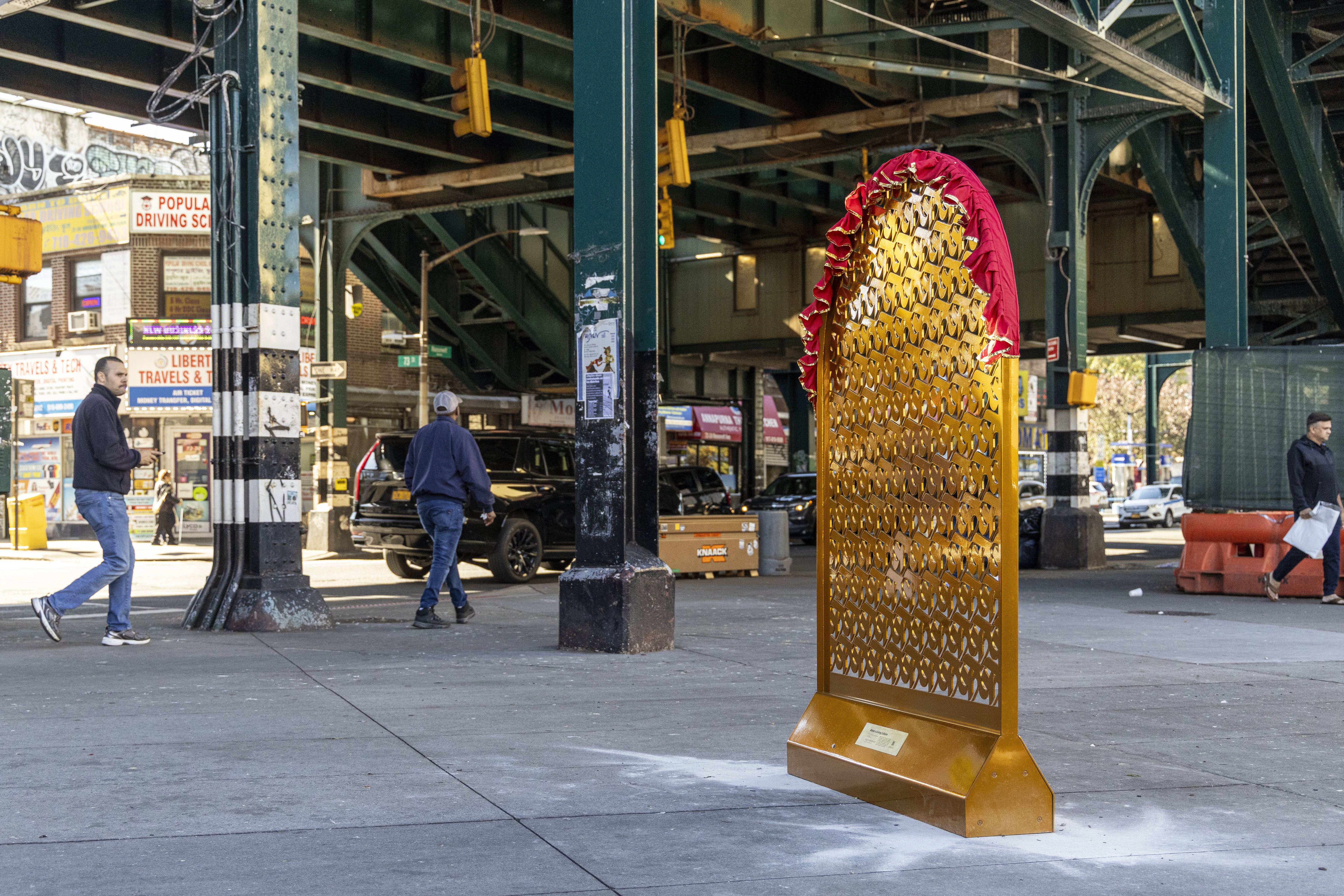 A tall gold sculpture with red fabric draped on top of it, is on display in a public plaza next to an elevated train station in Queens.