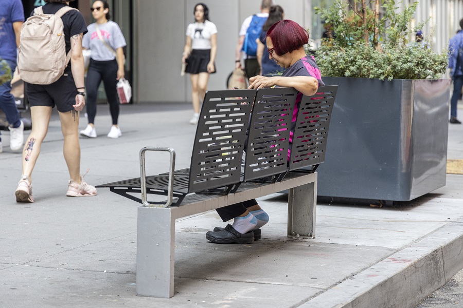 An older adult sits at a backed bench on a city sidewalk.