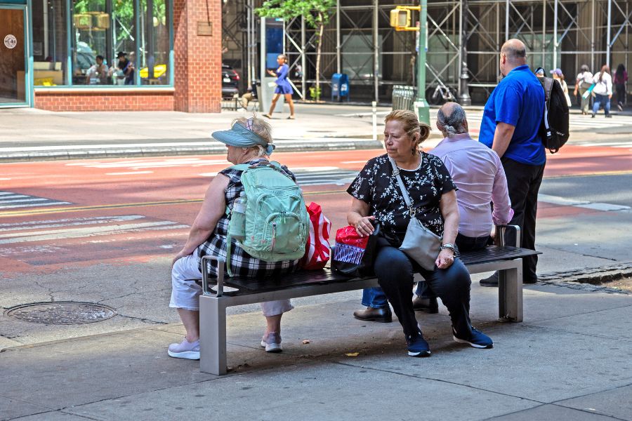 A woman sits on a backless bench as an M T A bus drives by.