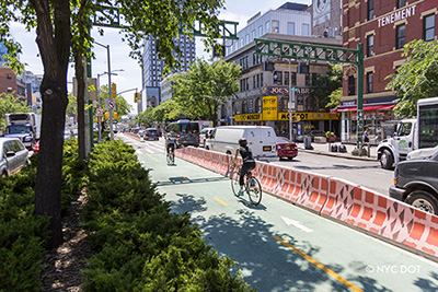 Two cyclists ride along a bike lane protected by a concrete jersey barrier painted with a red and pink design