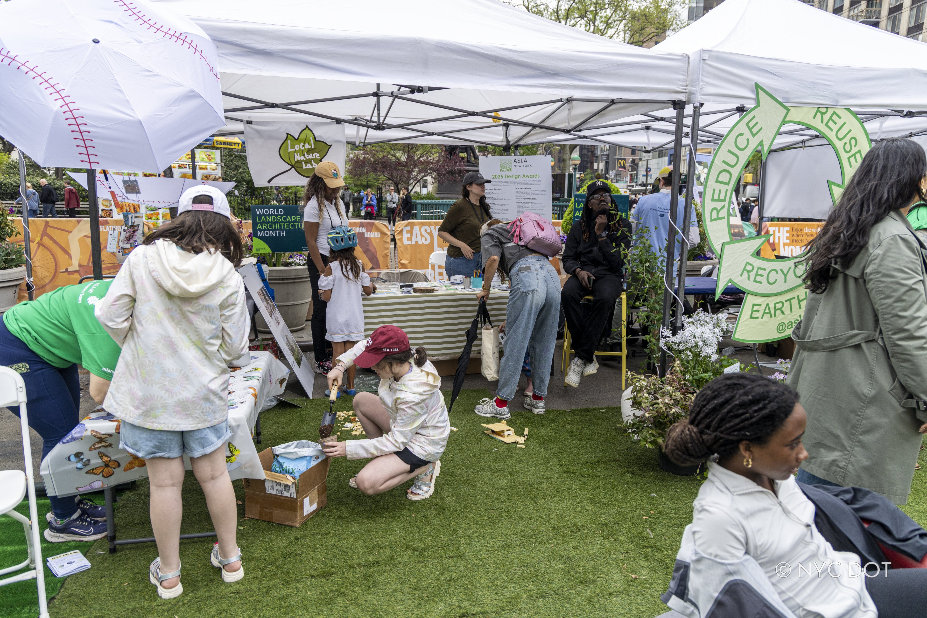 A large Reduce Reuse Recycle sign is attached to white tents setup on a car-free street while a local organization provides environmental activities for people in NYC.