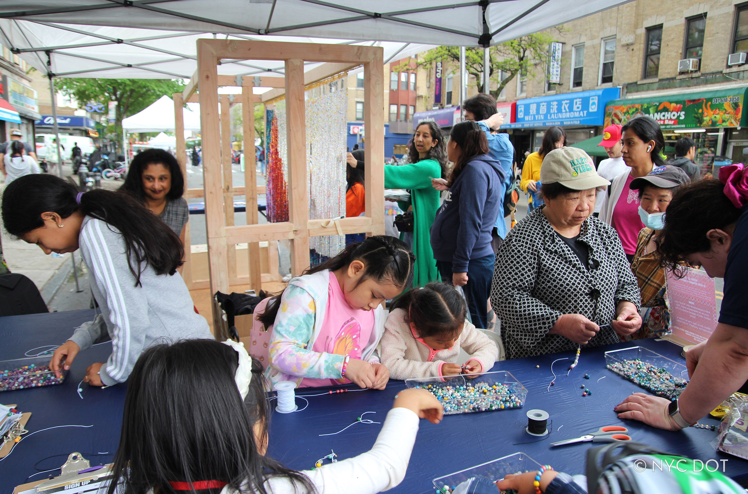 Children and adults make bead jewelry at a table setup next to an art installation featuring beadwork, presented during a car-free rainy event in Brooklyn.