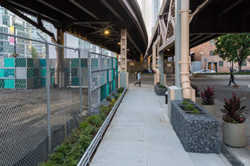 Planters line a clean sidewalk below an elevated roadway. One side of the sidewalk features a chain link fence and the other side features larger planters with green and red plants.