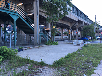 A concrete slab surrounded by green weeds at the base of a staircase from an elevated train.