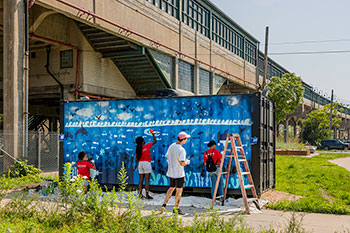 On a sunny day, volunteers paint a mural onto a shipping container positioned near a sidewalk adjacent to an elevated train station.