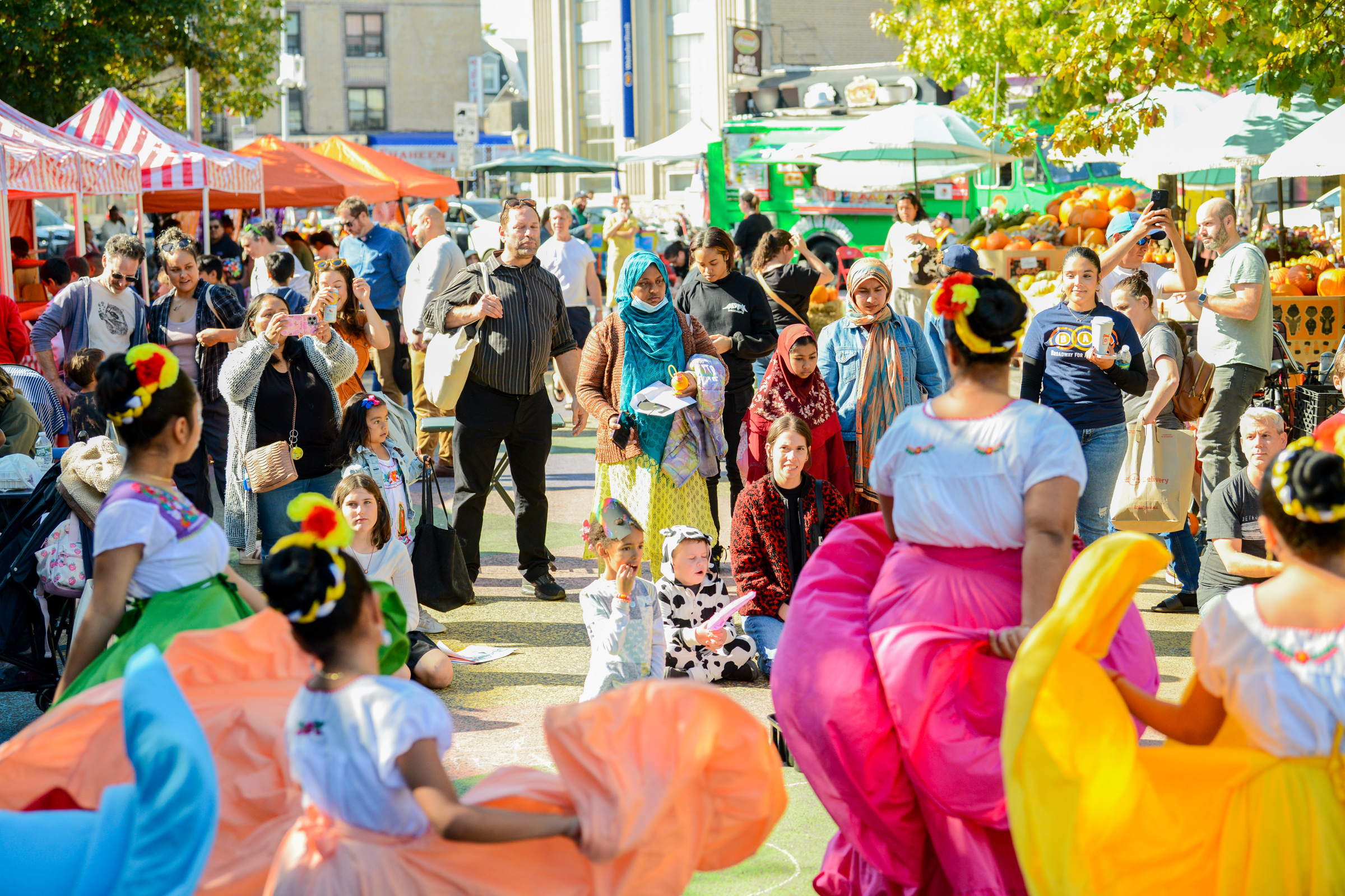 Young dancers in colorful skirts perform in front of a crowd at a plaza event.