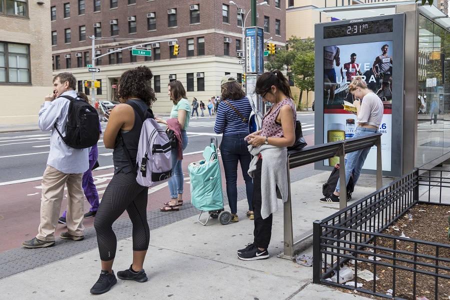 Two people lean against a leaning bar at a bus stop.