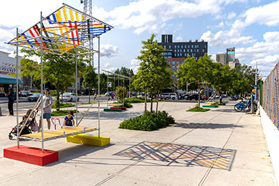 A child walks up to a artistic seating structure with a colorful overhead awning, set up with two other similar structures in a public plaza in Brooklyn.