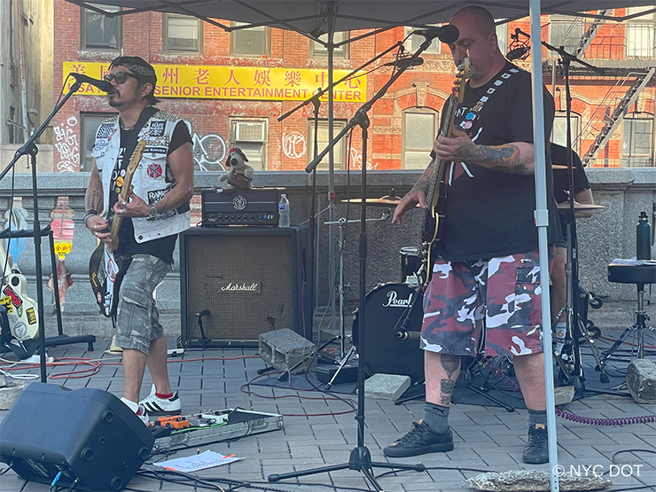 Two musicians play guitars and sing under a pop up tent in a plaza.