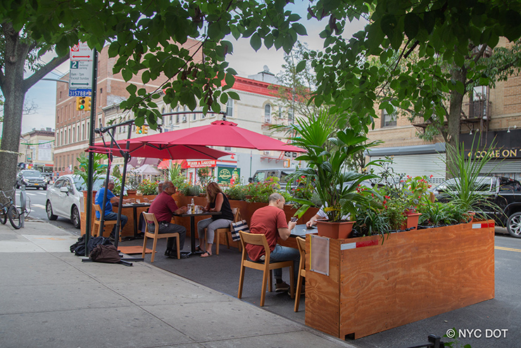 : People sit in an outdoor dining area set up in a roadway along the curb surrounded by a low barrier filled with plants. Large red umbrellas are positioned over two of the tables.