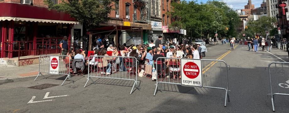 Multiple people enjoy outdoor seating on a street closed to vehicular traffic. In the background more people walk and bike on the same street.