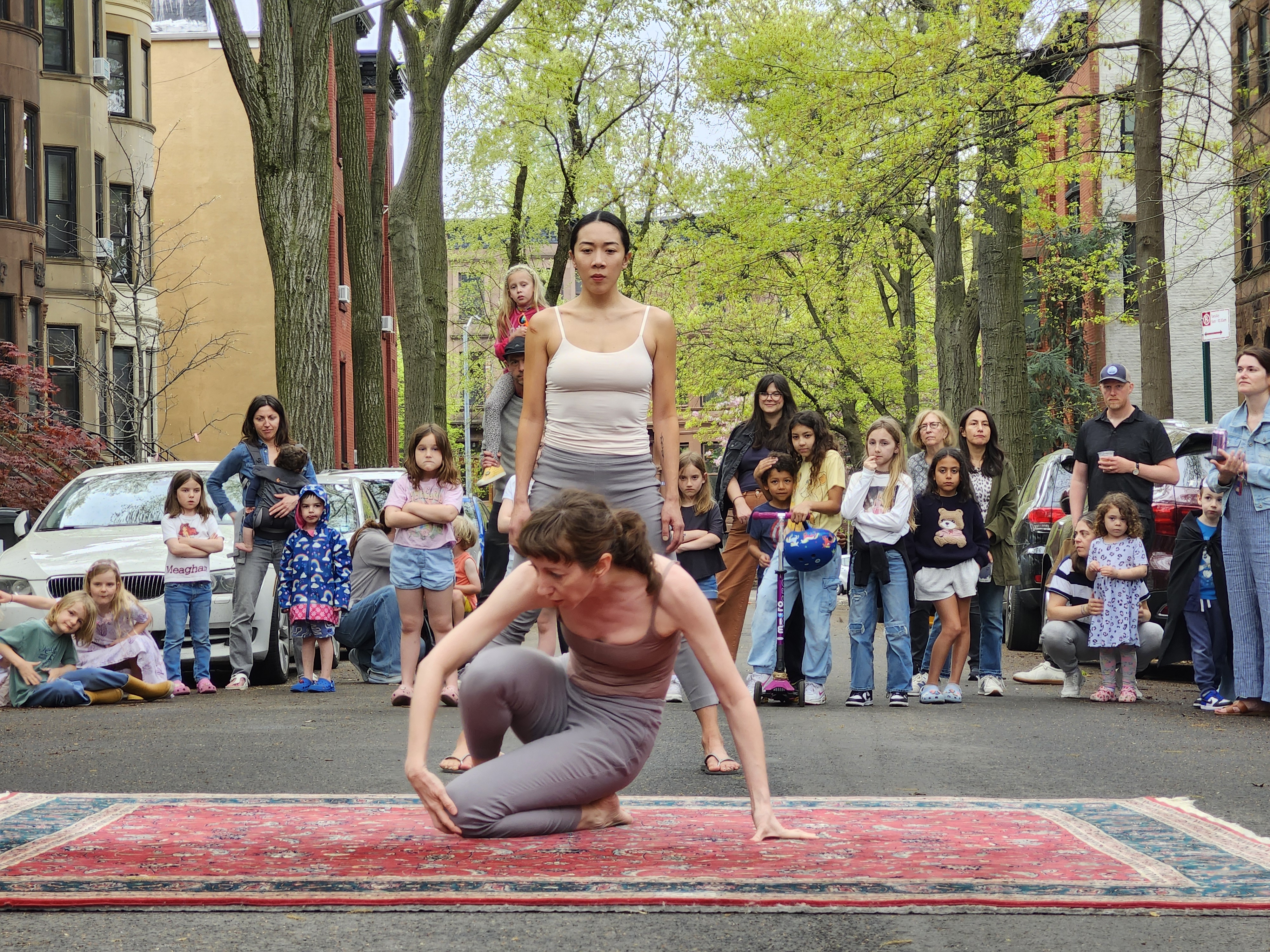 Two performers present interpretive dance on top of a carpet in the street while an engaged crowd of onlookers observes.