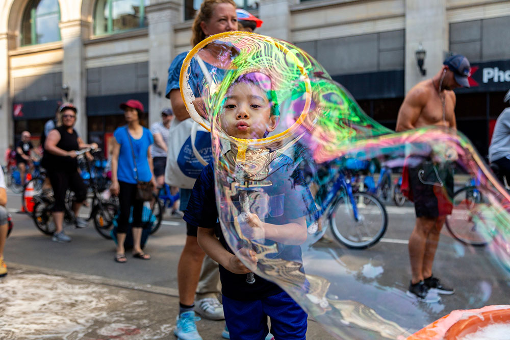 A young child looks through a large bubble while they play on a car-free street in New York City on a summer day.