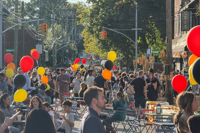 People walk and sit at tables and chairs with black, orange, red and yellow balloons setup on a street closed to traffic on Staten Island