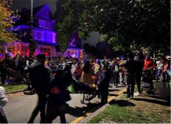 At night, large crowds of people dressed in costumes walk along a street closed to traffic to admire a large house covered in spooky lights and decorations for Halloween.