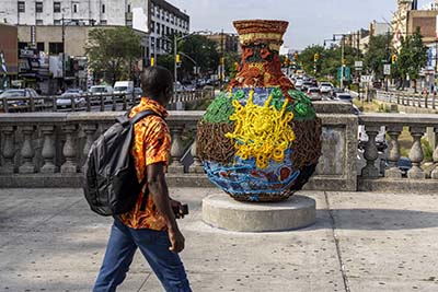 A man walks by and looks at a large water vessel sculpture covered in bright fibers with images of a yellow sun, brown and green mangrove trees, blue water and red fish.