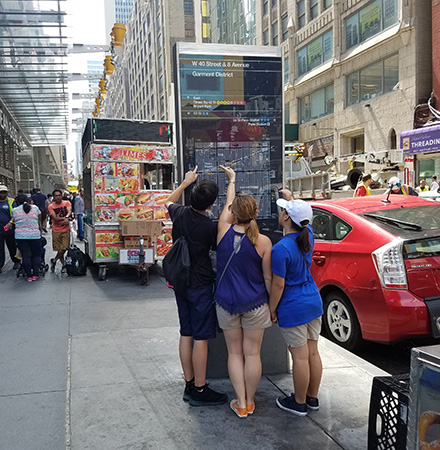 On a wide sidewalk, a group of people look at a tall sign maps on it