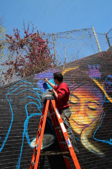 Artist standing at top of ladder painting a mural on a brick wall
