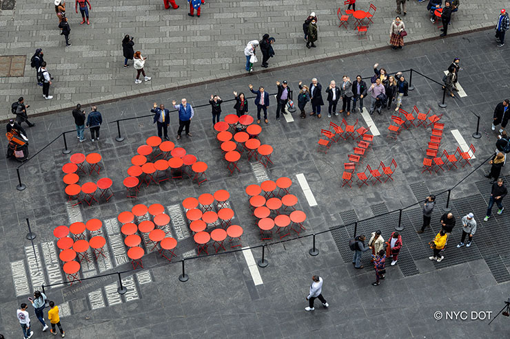 Aerial photo of red tables and chairs arranged to spell Car Free 22. N Y C D O T Commissioner Ydanis Rodriguez and other  N Y C D O T staff stand next to the arrangement and wave at the camera.
