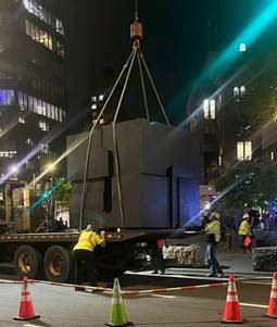Late at night a large black cube is lifted by crane onto the back of a truck.