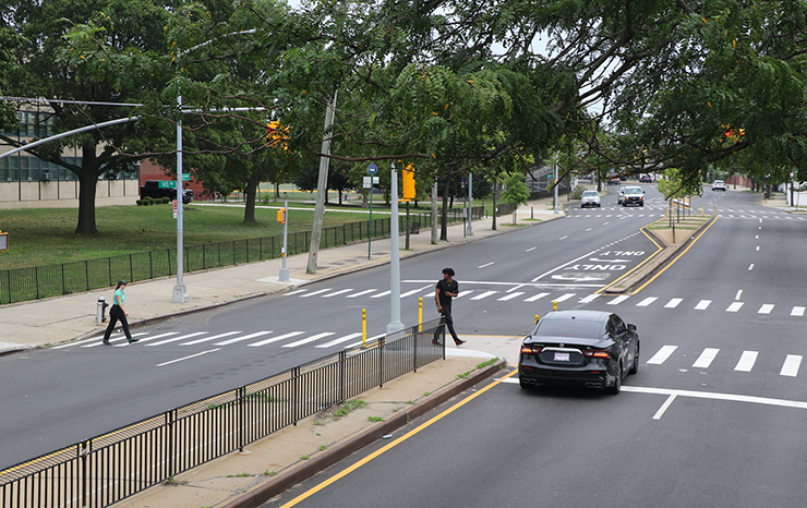 People walk in a crosswalk in front of a high school in Queens where there’s a new traffic signal controlling traffic. A black car is stopped at the red light.