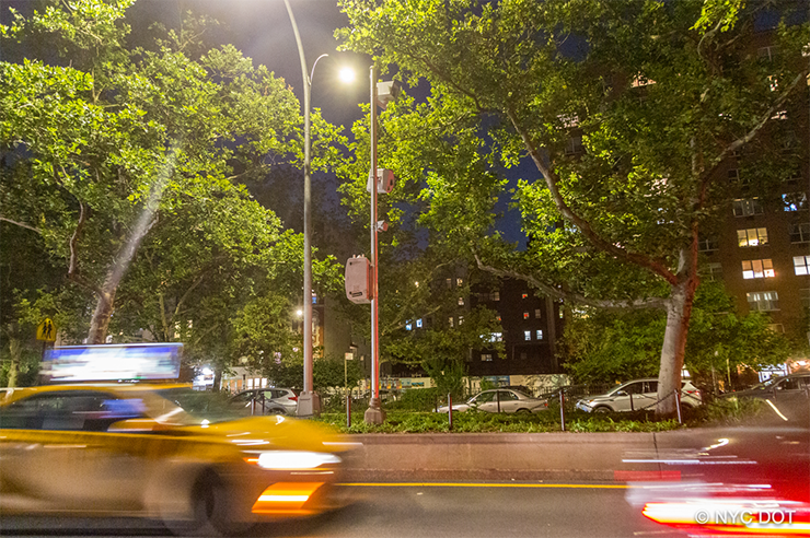 At night cars drive on a street in Manhattan past a tall pole with a speed camera attached to it
