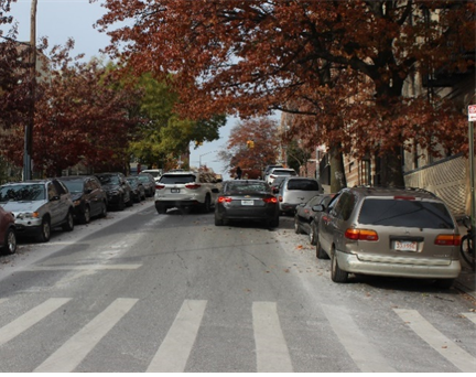 A driver passes a double parked car on a narrow two way street.