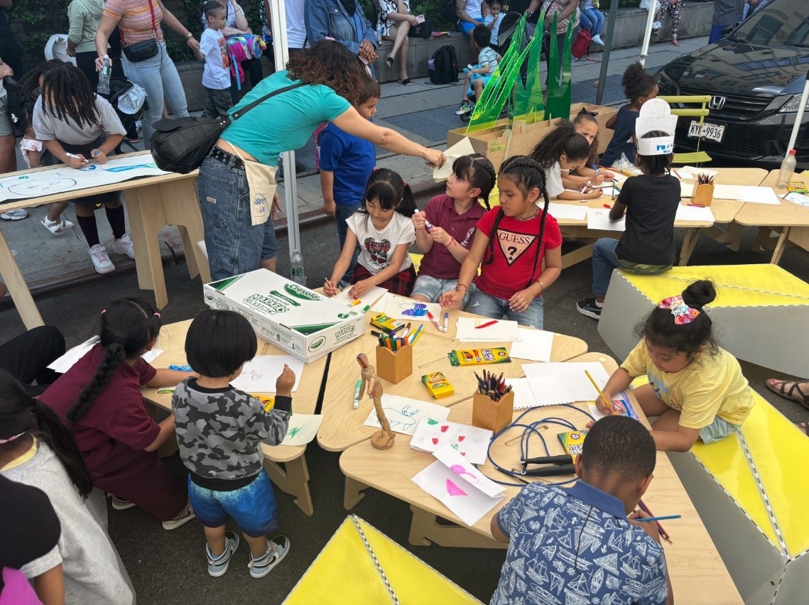 Children gather around moveable tables and chairs and bookcases on a street closed to traffic and open for student’s learning.