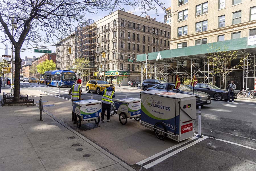 Two delivery workers place packages in cargo bikes parked in a curb extension along a busy roadway on the Upper West Side of Manhattan.