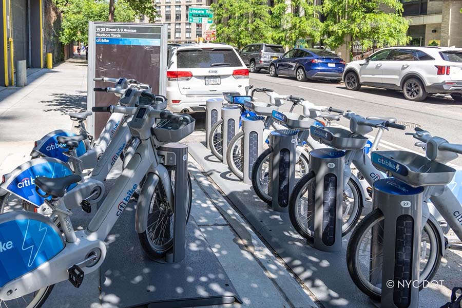 Two rows of gray Citi Bike docks filled with gray electric bikes. One dock is on a wide sidewalk and the second dock is on the roadway along the curb.