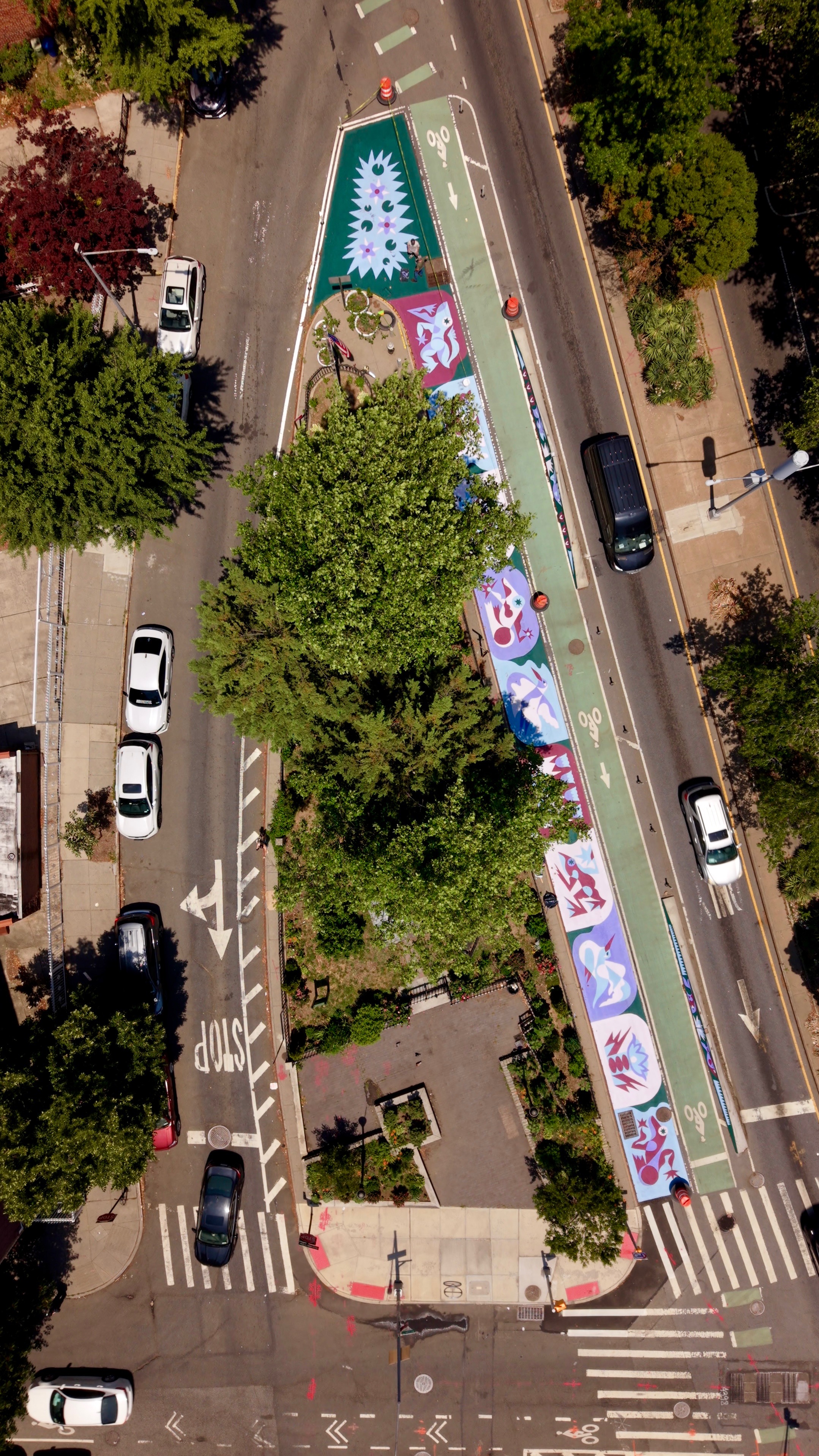 An aerial view of a street with a triangle island in the center one leg of the triangle has expanded pedestrian space decorated with an mural in greens, maroons and purple designs, next to a green bike lane.