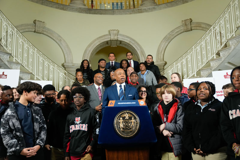 mayor adam is speaking at podium while a group of kids encircle behind him