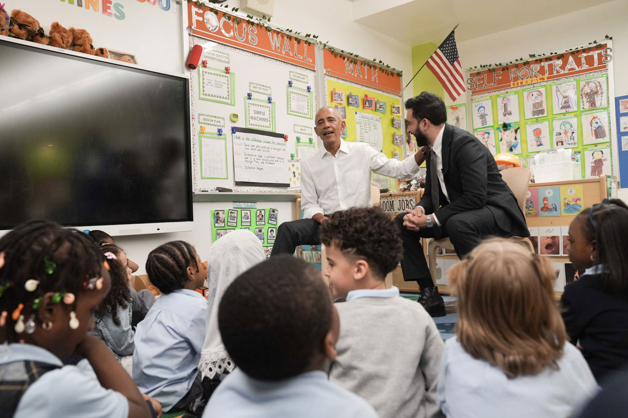 Former President Obama has his left hand rest on Mayor Mamdani's right shoulder, while a group kids sit in front of them.